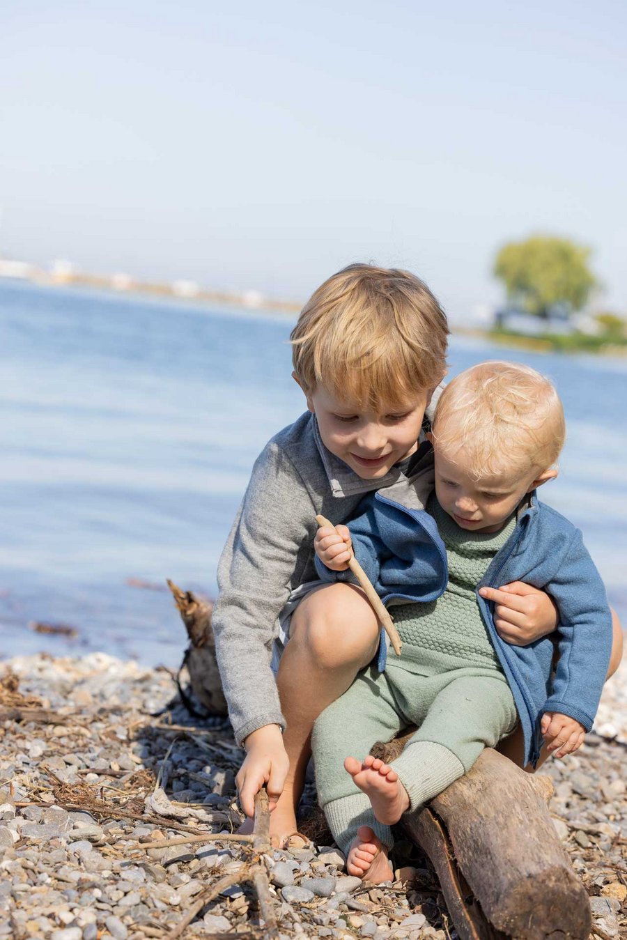 2 Kinder am Seeufer tragen disana Zipper-Jacken in Taubenblau & Grau, spielen glücklich auf einem Baumstamm. Sommer-Wollwalk