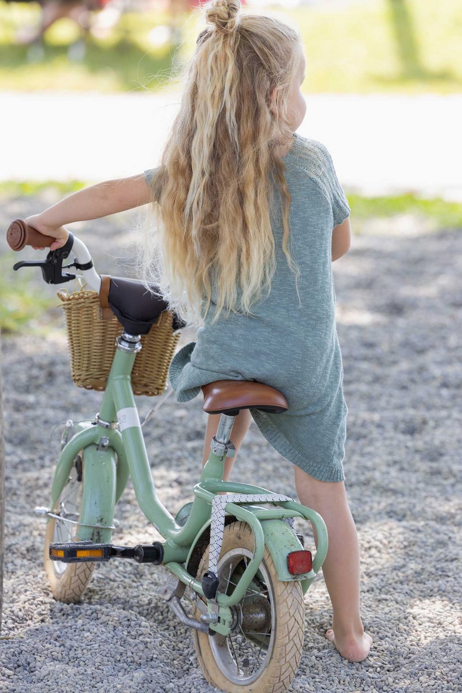 Ein Mädchen mit langen blonden Haaren im disana Leinen-Kleid Jade sitzt mit dem Rücken zur Kamera auf einem hellgrünen Rad