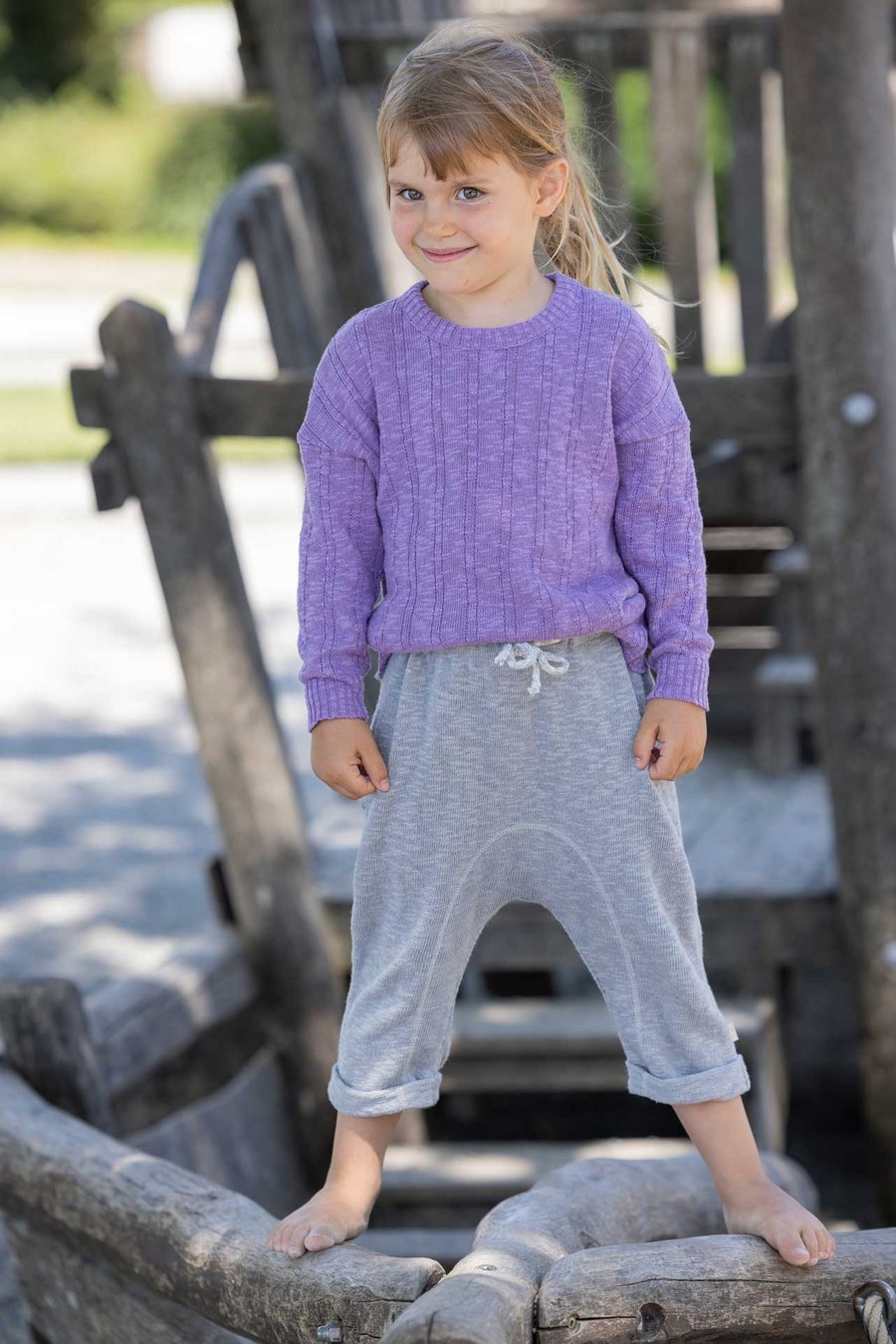 Mädchen mit goldbraunen Haaren trägt disana Leinen-Pullover, Flieder & Leinen-Strickhose, Grau stehend auf Holz-Klettergerüst