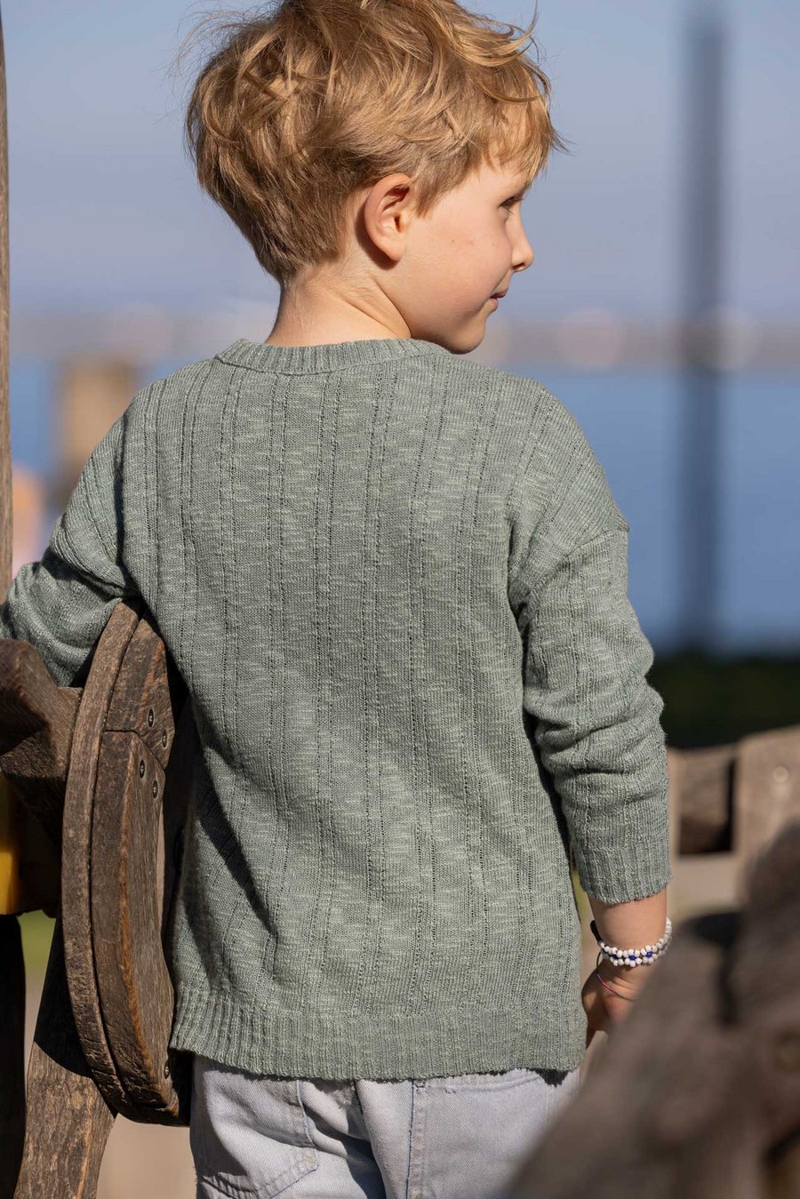 Blonder Junge im disana Leinen-Pullover, Jade, auf Holz-Klettergerüst, Rückenansicht, draußen, Hintergrund blauer Himmel