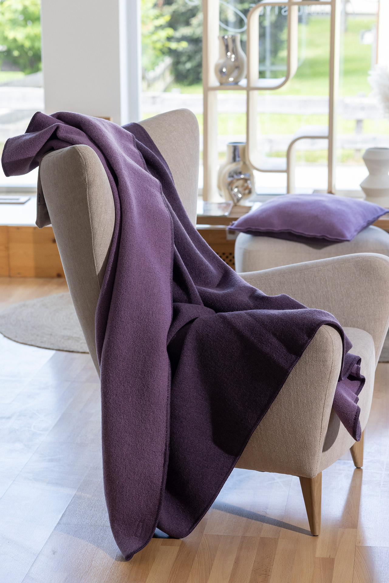 disana Doubleface-Decke in Aubergine-Flieder auf einem beigen Sessel drapiert in einem hellen Raum, Wollwalk: Bio-Merinowolle