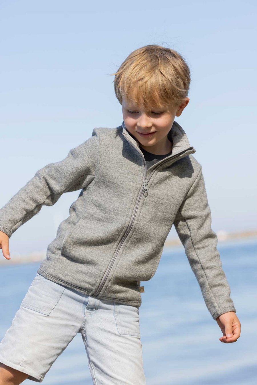 Kind in grauer disana Zipperjacke aus Bio-Merino-Wolle steht am Strand eines Seeufers