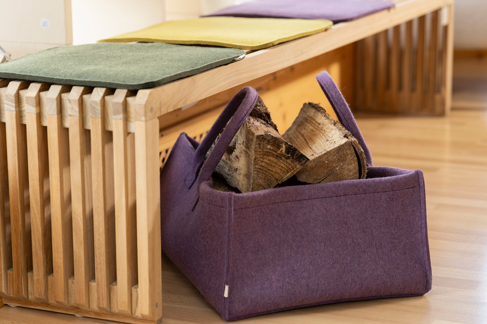 disana Filz-Aufbewahrungskorb in Aubergine mit Holzscheiteln gefüllt vor einer Holz Lamellenbank mit bunten Filz-Auflagen.