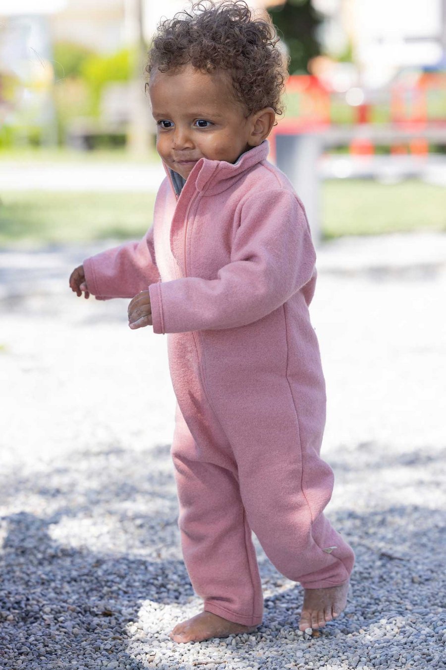 Dunkelhäutiges Kind im disana Walk-Anzug Softpinkt läuft barfuß auf Kieselsteine, Sommer-Wollwalk 260g/m² aus Bio-Merinowolle