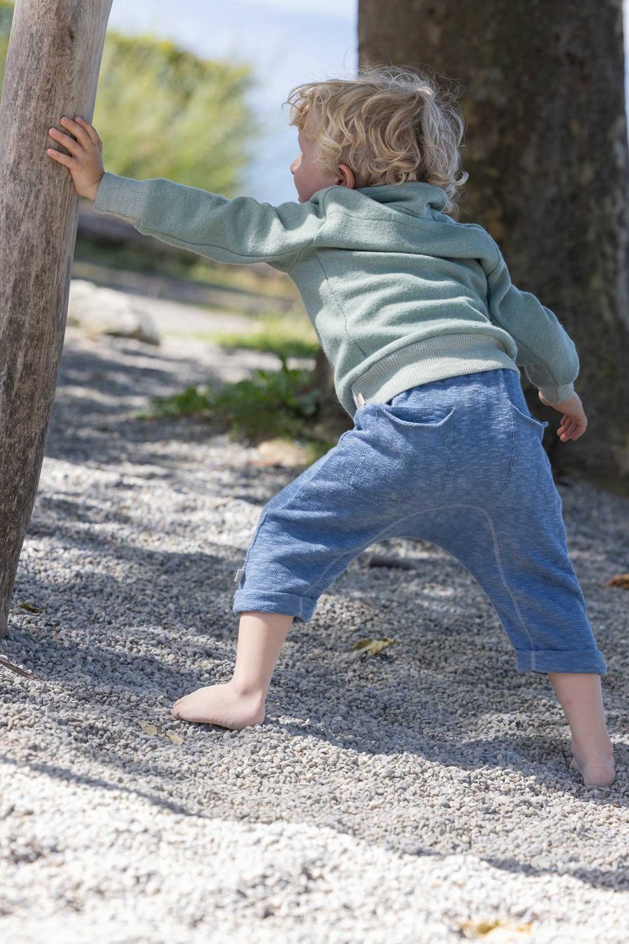 Kind in disana Troyer, Jade und Leinen-Strickhose Taubenblau hält sich mit der linken Hand an einem Stamm, Rückenansicht
