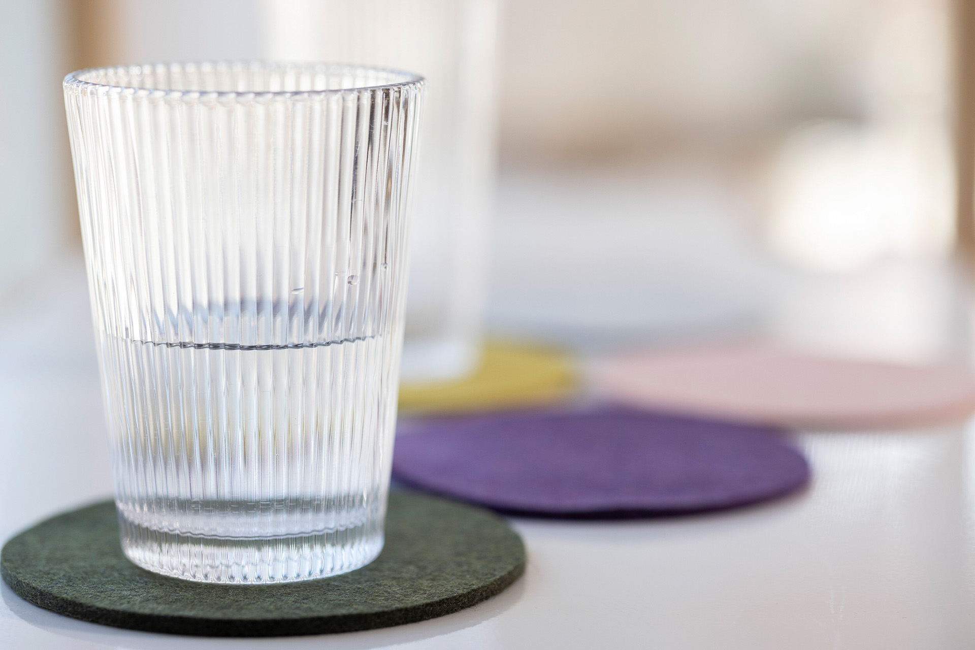 Geriffeltes Wasserglas auf olivem disana Filz-Untersetzer; im Hintergrund weitere bunte Untersetzer aus Wollfilz.