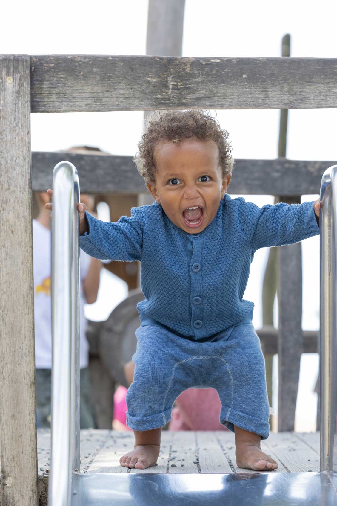 Baby wearing a dove blue honeycomb cardigan by disana on slide, enjoying outdoor play - Spring/Summer collection