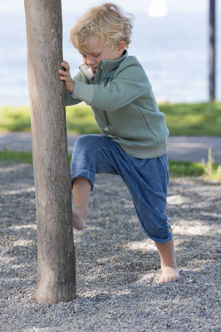 Blonder Junge im disana Troyer in Jade und Leinen-Strickhose in Taubenblau versucht an einem Baumstamm zu klettern