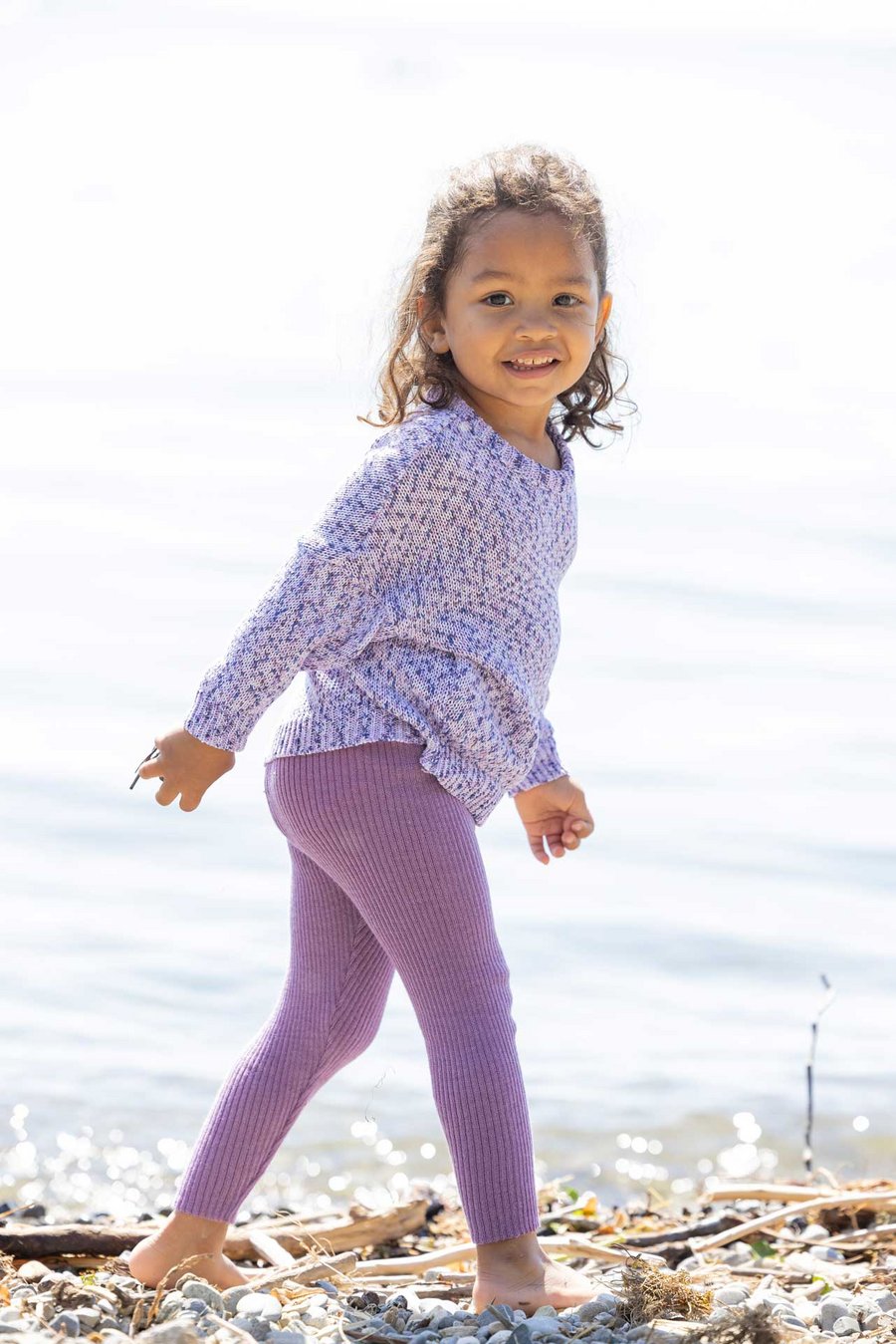 Kind hellbrauner Haut in disana Baumwollpulli Spray Leggings-Light Flieder am Seeufer läuft nach rechts lächelt in die Kamera