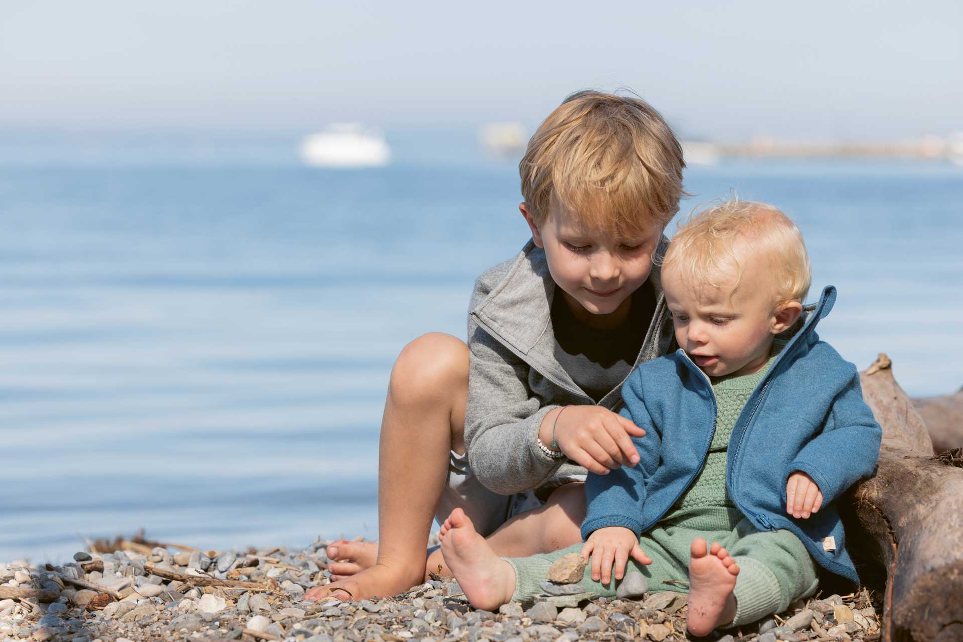 2 Kinder am Strand in disana Zipper-Jacken in Grau & Taubenblau, kombiniert mit Pumphose & Wabenstrick-Pullover in Jade