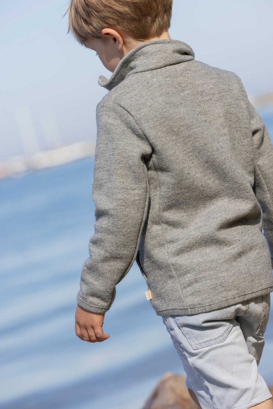 Blondes Kind in grauer disana Zipper-Jacke am Seeufer, dreht den Rücken zur Kamera - Wollwalk, 260 g/m² aus Bio-Merinowolle