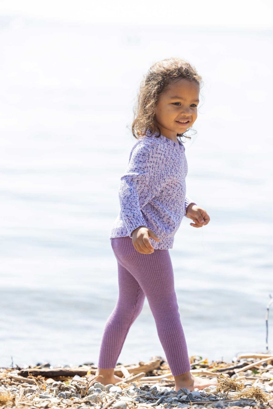 Kind hellbrauner Haut in disana Baumwollpulli Spray, Leggings-Light Flieder am Seeufer läuft nach rechts schaut in die Kamera