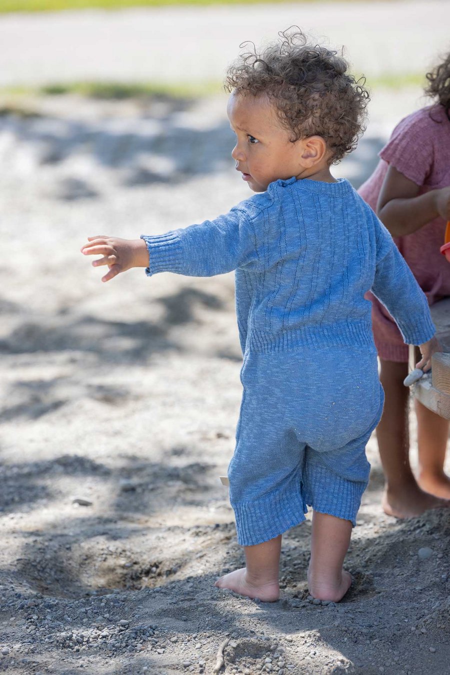 Kleinkind trägt disana Leinen-Strampler, Taubenblau, dreht der Kamera den Rücken zu & schaut nach links, barfuß im Sand