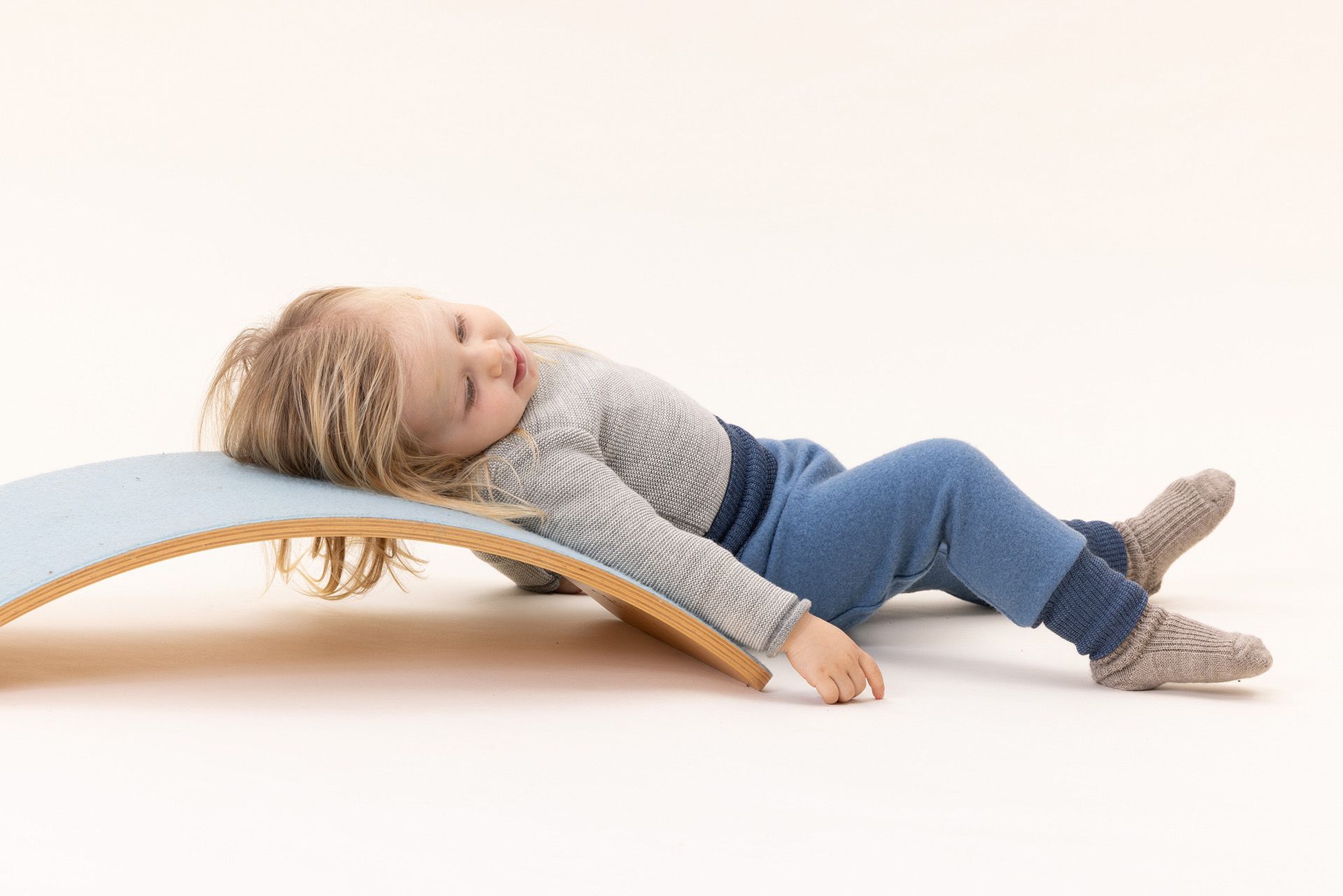 Blondes Kind liegt auf Balanceboard in disana Pumphose Taubenblau, Melange-Pullover Grau-Natur, beiger Hintergrund