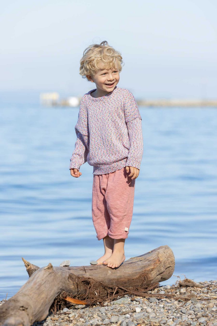 Blonder Junge in disana Baumwoll-Pullover "Spray" Softpink Spray & Leinen-Strickhose Softpink, steht auf Holzstamm am Seeufer