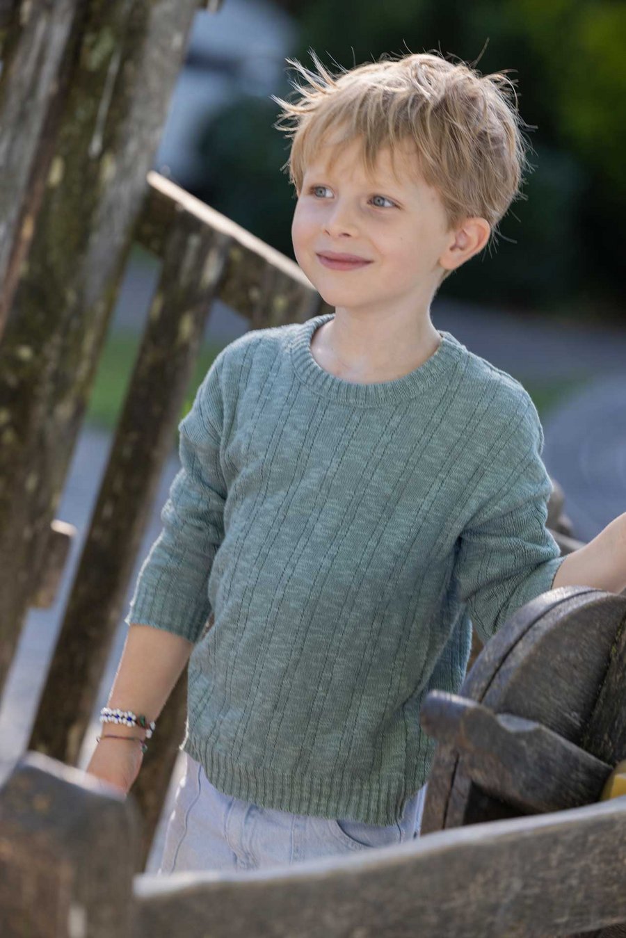 Blonder Junge trägt disana Leinen-Pullover in Jade spielt mit einem verwitterten Holzrad. Pullover Bio-Baumwolle & Bio-Leinen